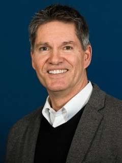 Portr&auml;t eines Mannes mit grauen Haaren, l&auml;chelnd, in grauem Blazer und wei&szlig;em Hemd vor blauem Hintergrund.