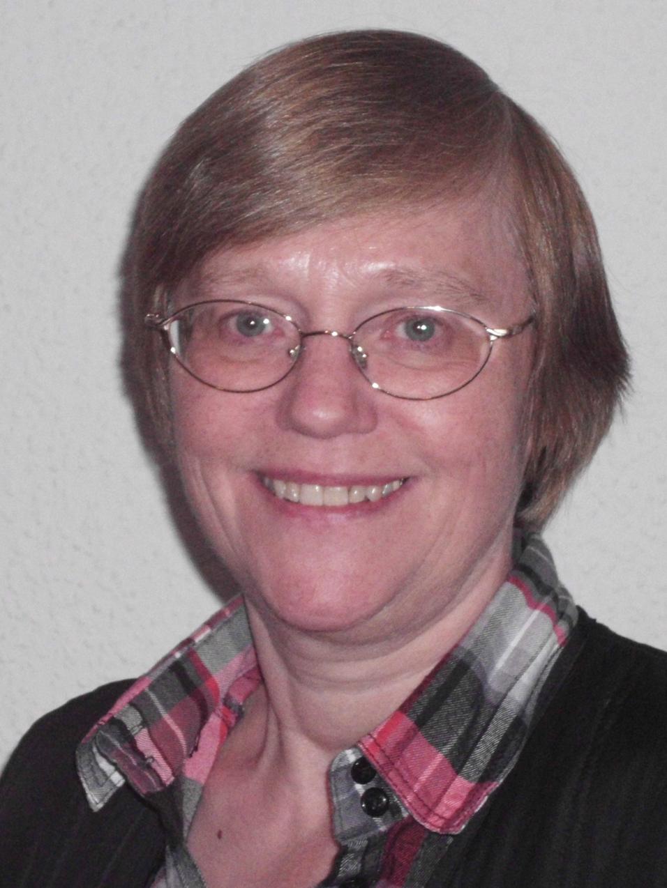 Portr&auml;t einer l&auml;chelnden Frau mit kurzen, braunen Haaren und Brille, vor einer hellen Wand. Sie tr&auml;gt ein kariertes Hemd.