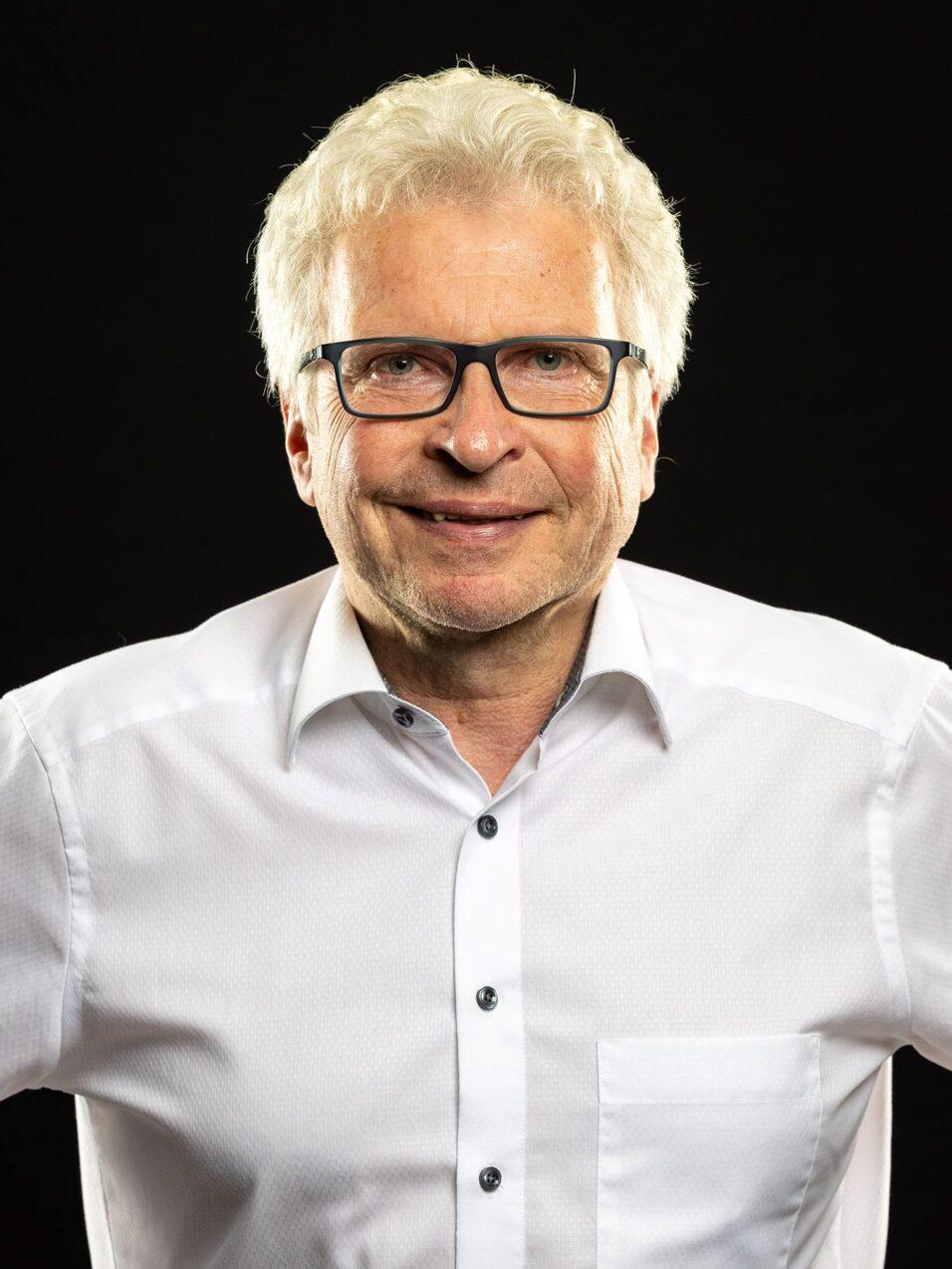 Mann mit Brille und kurzem, grauem Haar in wei&szlig;em Hemd vor schwarzem Hintergrund.