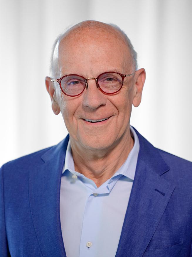 &Auml;lterer Mann mit Brille und Anzug l&auml;chelt vor einem hellen, neutralen Hintergrund.