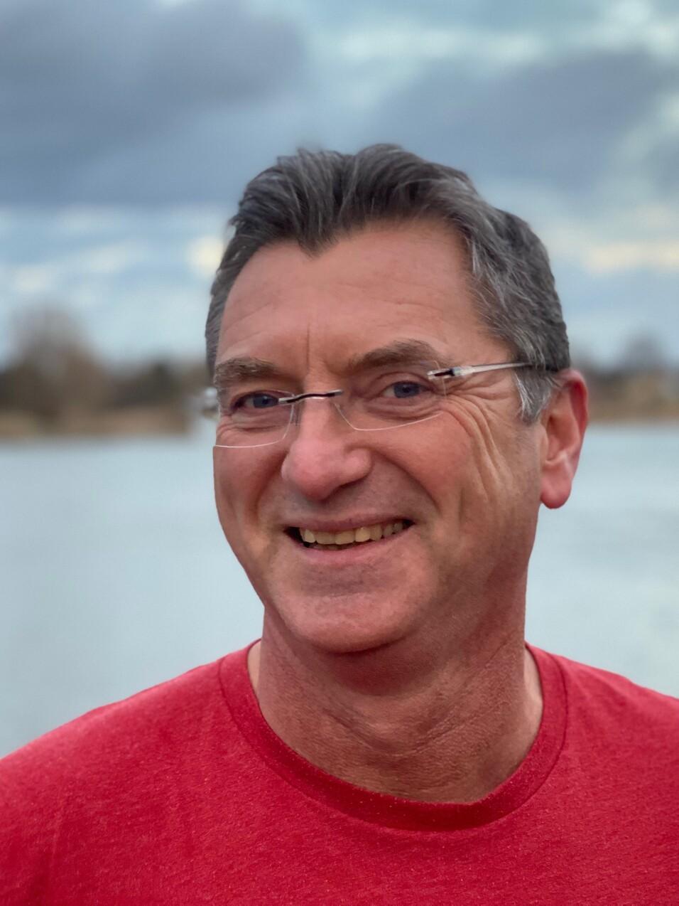 L&auml;chelnder Mann mit Brille tr&auml;gt ein rotes T-Shirt, vor einem Gew&auml;sser mit bew&ouml;lktem Himmel.