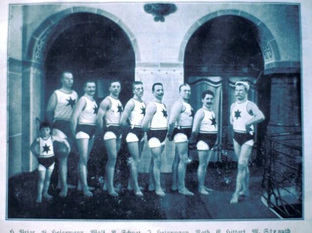 Schwarz-wei&szlig; Foto von M&auml;nnern in Badeanz&uuml;gen mit Sternen, aufgestellt in einer Halle mit B&ouml;gen.