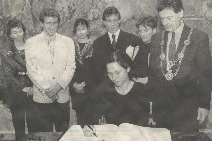 Frau in schwarzer Kleidung unterschreibt in ein gro&szlig;es Buch, umgeben von einer Gruppe von Menschen in formeller Kleidung.