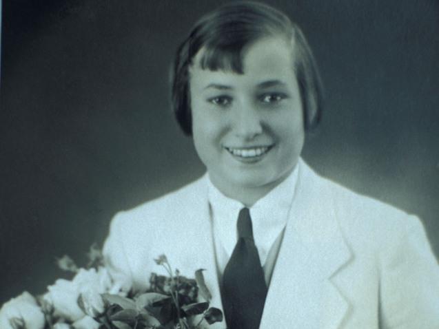 Junge Frau im wei&szlig;en Anzug mit Kravatte, l&auml;chelnd, h&auml;lt einen Blumenstrau&szlig; und eine Fahne mit olympischen Ringen.