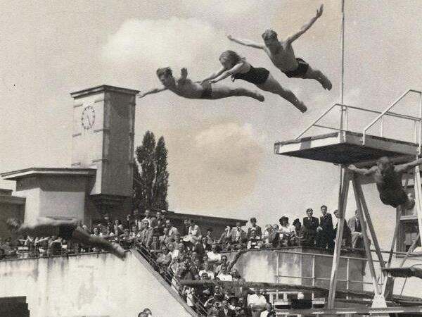 Gruppe von Kindern springt vom Sprungbrett ins Wasser, w&auml;hrend Zuschauer am Rand eines Schwimmbeckens zuschauen.