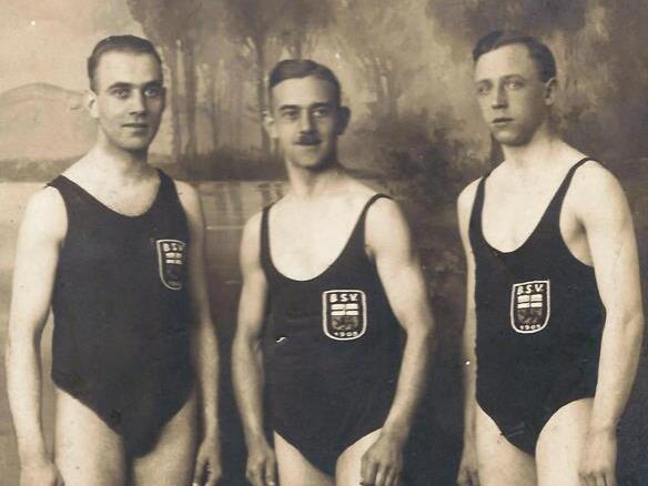 Drei M&auml;nner im Badeanzug stehen vor einer verschwommenen Landschaft, jeweils mit einem Aufn&auml;her des BSV.