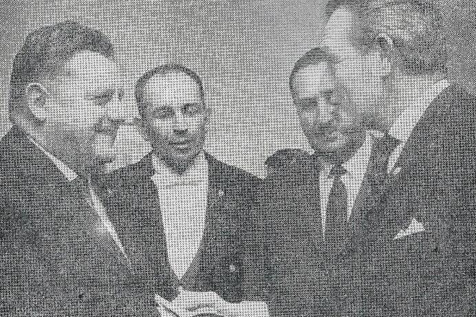 Vier M&auml;nner stehen in einem Raum, einer &uuml;berreicht einem anderen ein Dokument. Alle wirken an einem Gespr&auml;ch beteiligt.