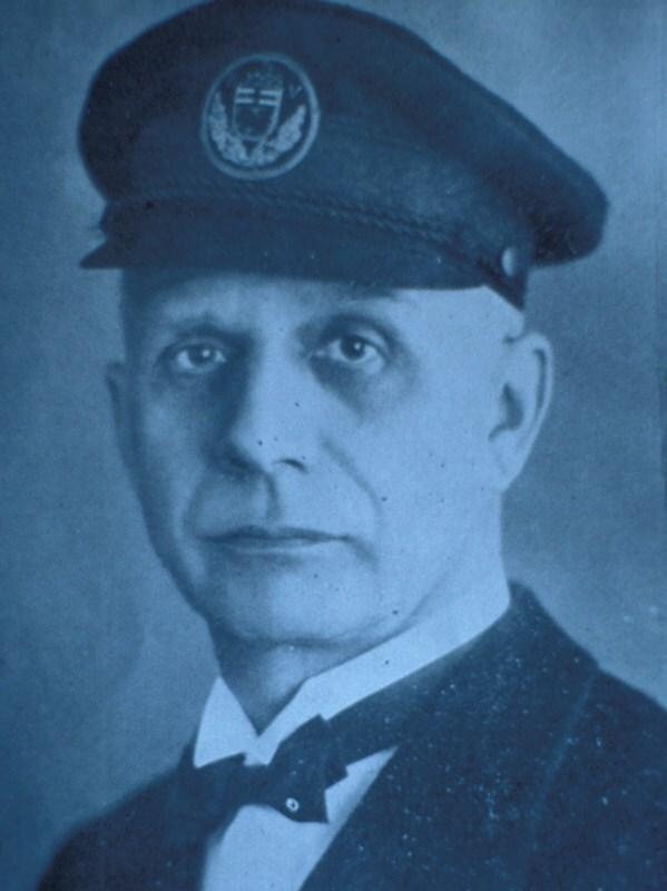 Schwarzwei&szlig;portr&auml;t eines Mannes in Uniform mit M&uuml;tze und Fliege, ernsthaftem Ausdruck auf bluet&ouml;nigem Hintergrund.