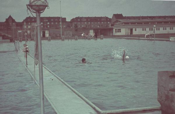 Schwimmer in einem Freibad, das von einem Steg umgeben ist; im Hintergrund sind Geb&auml;ude zu sehen.