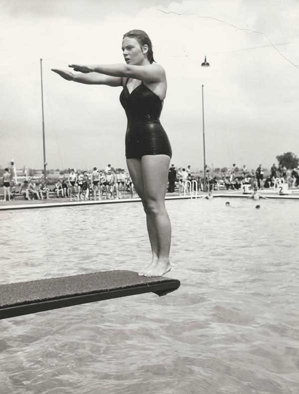 Springerin auf einem Sprungbrett, bereit zum Sprung ins Schwimmbecken, umgeben von Zuschauern.