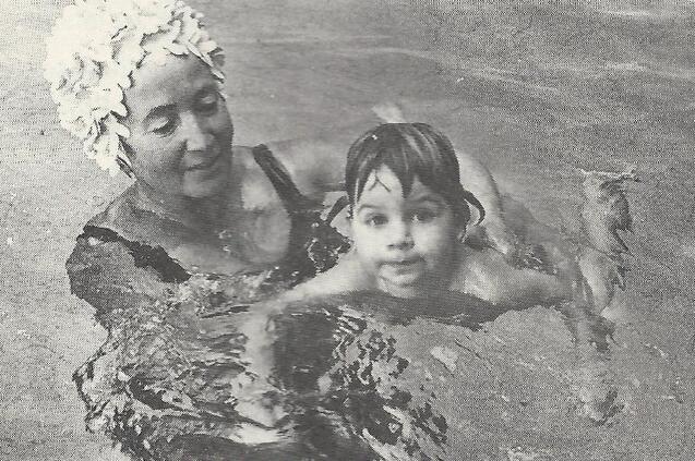 &Auml;ltere Frau mit Badekappe h&auml;lt ein schwimmendes Kind im Wasser. Beide l&auml;cheln in einer entspannten Badestimmung.