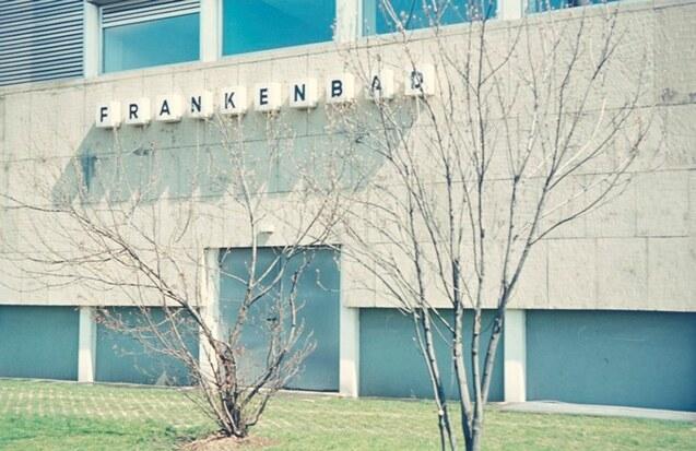 Fassade des Frankenbads mit gro&szlig;en Buchstaben, grauem Betonsockel und kahlbl&auml;ttrigem Baum im Vordergrund.