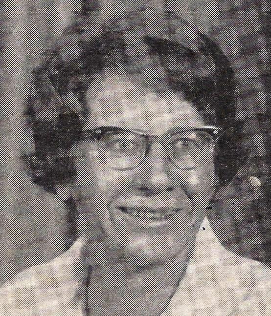 Schwarz-Wei&szlig;-Foto einer l&auml;chelnden Frau mit Brille und lockigem Haar, vor einem neutralen Hintergrund.