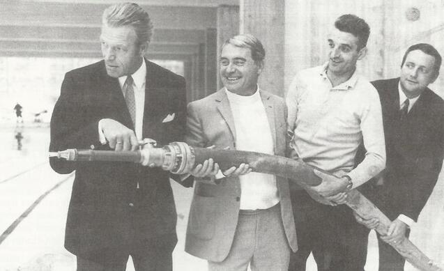 Vier M&auml;nner halten gemeinsam ein gro&szlig;es Rohr in einem modernen Raum mit Fenstern im Hintergrund.