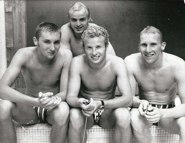 Vier junge M&auml;nner mit nacktem Oberk&ouml;rper posieren l&auml;chelnd neben einem Schwimmbecken. Sie tragen Badehosen und halten Gegenst&auml;nde in den H&auml;nden.