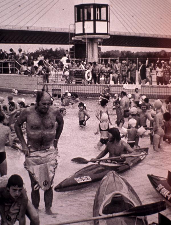 Menschen schwimmen und spielen im Wasser eines Freibades, mehrere Kinder und Kajaks im Vordergrund. Hochhaus im Hintergrund.
