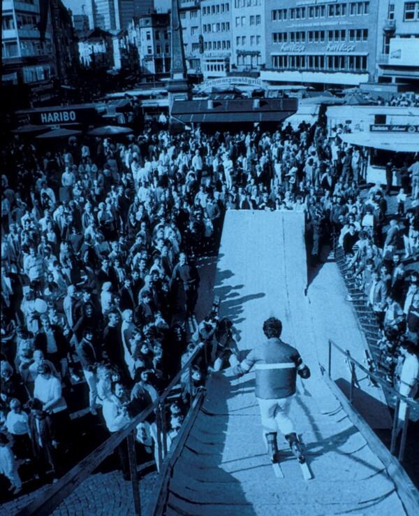 Skifahrer springt von einer Holzrampe vor einer gro&szlig;en Menschenmenge in einer urbanen Umgebung.