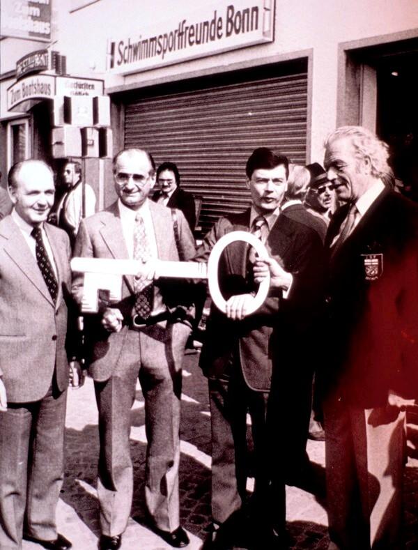 Vier M&auml;nner stehen vor einem Geb&auml;ude mit dem Schild &bdquo;Schwimmsportfreunde Bonn&ldquo; und halten einen gro&szlig;en Schl&uuml;ssel.