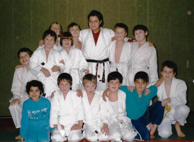 Gruppe von Kindern in wei&szlig;en Judogis und einem Trainer in Schwarz, l&auml;chelnd auf einer Judomatte posierend.