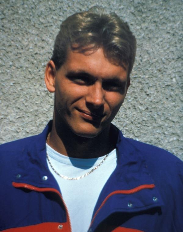 Portr&auml;t eines jungen Mannes mit kurzer, blonder Frisur, blau-rotem Windbreaker und Kette vor einer grauen Wand.