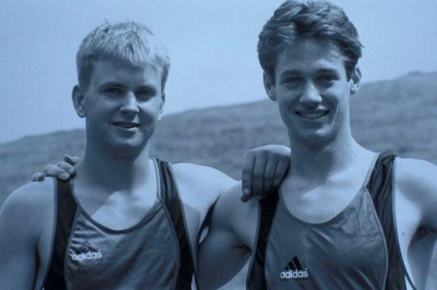 Zwei junge Sportler mit kurzen Haaren in Sportanz&uuml;gen stehen nebeneinander und l&auml;cheln freundlich in die Kamera.