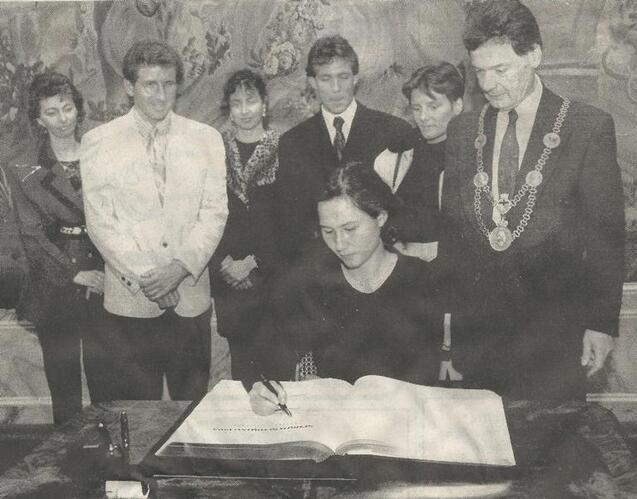 Frau in schwarzer Kleidung unterschreibt in ein gro&szlig;es Buch, umgeben von einer Gruppe von Menschen in formeller Kleidung.