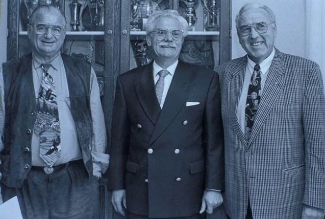 Drei &auml;ltere M&auml;nner in Anz&uuml;gen stehen l&auml;chelnd vor einem Schrank mit Pokalen und Troph&auml;en.