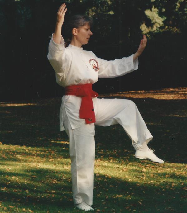 Frau in wei&szlig;em Karateanzug mit rotem G&uuml;rtel, die einen Karate-Kampfstand auf einer gr&uuml;nen Wiese einnimmt.