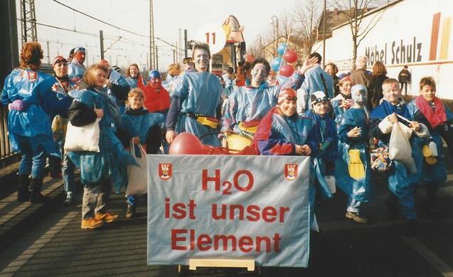 Gruppe von verkleideten Menschen in blauer Kleidung mit einem Banner "H₂O ist unser Element" bei einer Stra&szlig;enveranstaltung.