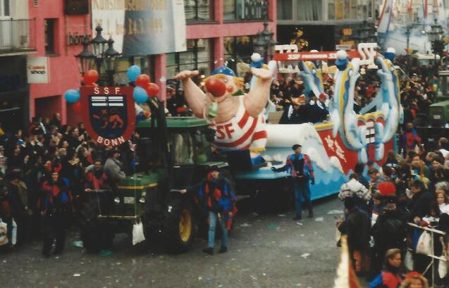 Bunte Karnevalswagen mit einer gro&szlig;en Figur und Menschen in festlicher Kleidung auf einer belebten Stra&szlig;e.