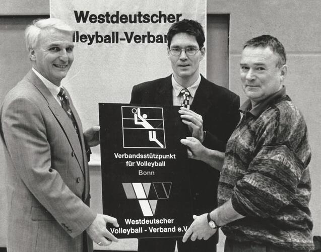 Drei M&auml;nner halten ein Schild mit Informationen zum Volleyballverband in Bonn vor einem Banner des Westdeutschen Volleyball-Verbandes.