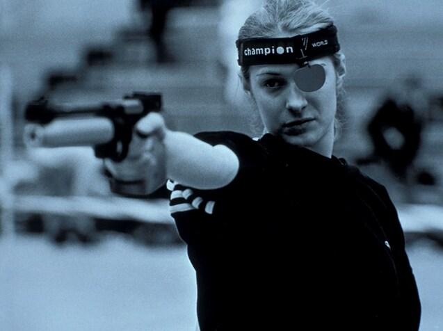 Sportsch&uuml;tzin mit schwarzem Shirt und Augenbinde zielt mit einer Pistole auf den Betrachter.