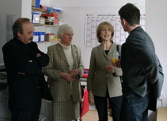 Vier Personen stehen in einem Raum und unterhalten sich, w&auml;hrend eine Frau ein Glas mit einem Getr&auml;nk h&auml;lt.