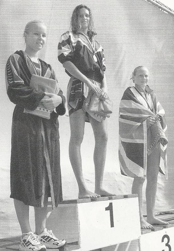 Drei Schwimmerinnen auf dem Siegerpodest nach einem Wettkampf, die erste h&auml;lt eine Medaille, die anderen tragen Handt&uuml;cher.