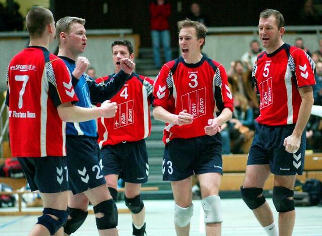 Sechs Volleyballspieler in roten Trikots jubeln und feiern im Sportgel&auml;nde. Zuschauer sind im Hintergrund sichtbar.
