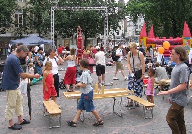 Bunte Menschenmenge auf einem Platz, Familien und Kinder interagieren, im Hintergrund ein Spielplatz und eine Performance.