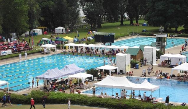 Blick auf ein Freibad mit Schwimmern, Zelten und St&auml;nden; zahlreiche Besucher am Rand der blauen Schwimmbecken.