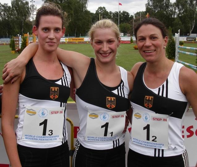 Drei Athletinnen in Sportkleidung stehen eng beieinander und l&auml;cheln vor einer Wettkampfst&auml;tte f&uuml;r Modernen F&uuml;nfkampf.