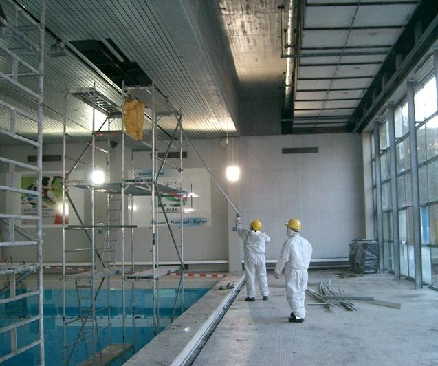 Zwei Arbeiter in Schutzkleidung und Helmen stehen vor einem unvollendeten Schwimmbad, umgeben von Ger&uuml;st und Baumaterial.