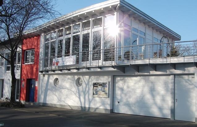 Moderne Geb&auml;ude mit gro&szlig;en Fenstern, roter Fassade und glattem Garagentor, vor blauem Himmel und Schatten von B&auml;umen.