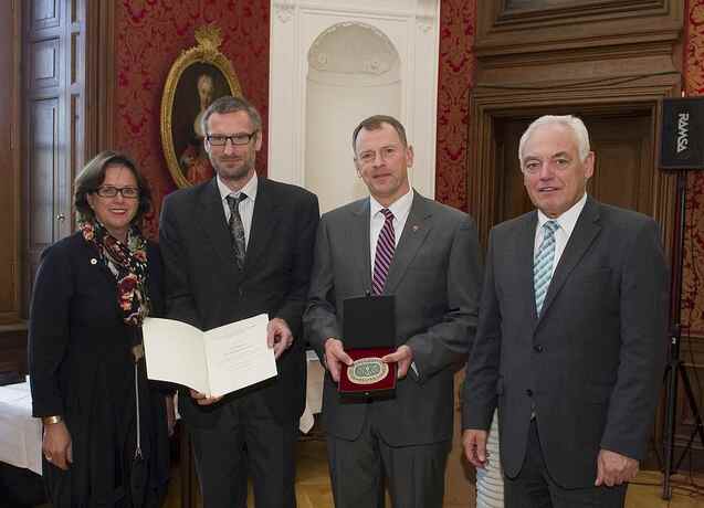 Vier Personen stehen in einem historischen Raum. Zwei halten Urkunde und Medaille, im Hintergrund ein Portr&auml;t.