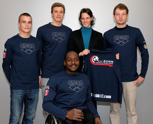 Vier Personen in blauen Sweatshirts stehen und sitzen gemeinsam vor einer grauen Wand, eine h&auml;lt ein T-Shirt mit Logo.