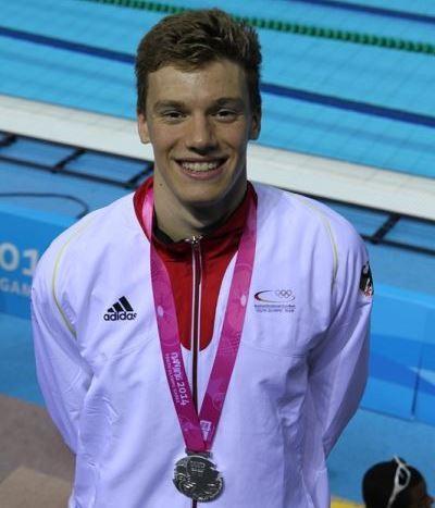 Junger Schwimmer mit Silbermedaille um den Hals, l&auml;chelt vor einem Schwimmbecken in Wettkampfumgebung.