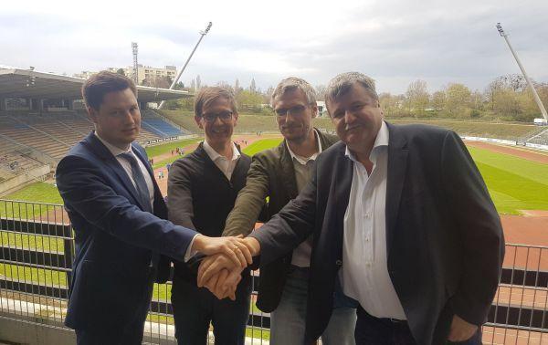 Vier M&auml;nner stehend auf einer Trib&uuml;ne, die H&auml;nde in der Mitte &uuml;bereinander gelegt, mit einem Sportstadion im Hintergrund.