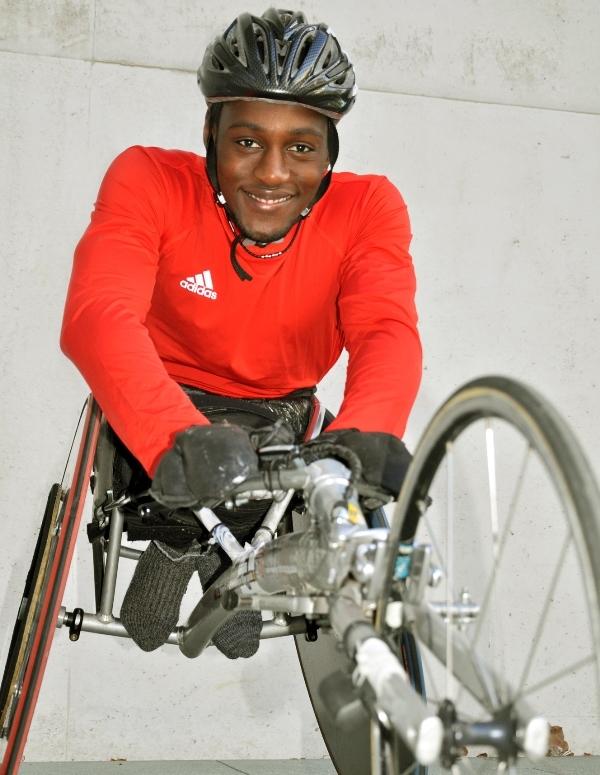 L&auml;chelnder Mann in rotem Shirt und Helm sitzt in einem Handbike und blickt kameraw&auml;rts. Hintergrund ist neutral.