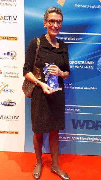 Frau in schwarzem Kleid h&auml;lt einen Preis, steht vor einer Bannerwand bei einer Preisverleihung.