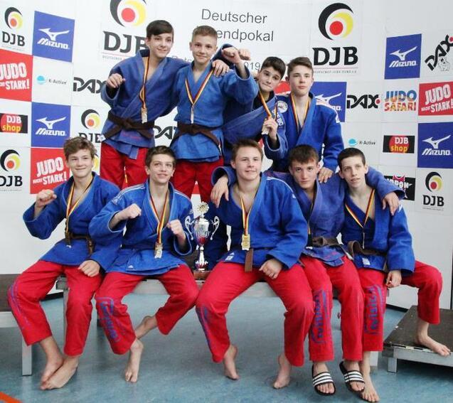 Gruppe von Judokas in blauen und roten Anz&uuml;gen mit Medaillen, posierend vor einem Pokal und Sportbanner.