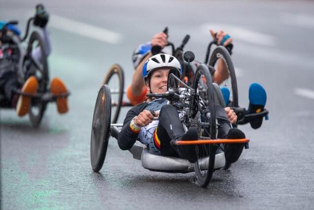 Handcyclerin f&auml;hrt auf nasser Stra&szlig;e, vorne im Bild l&auml;chelt sie, im Hintergrund weitere Sportler in Handbikes.