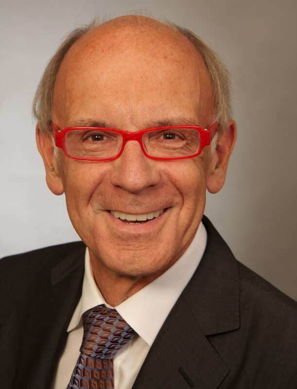 Portrait eines l&auml;chelnden Mannes mit roten Brillengestell, Anzug und Krawatte, vor einem neutralen Hintergrund.