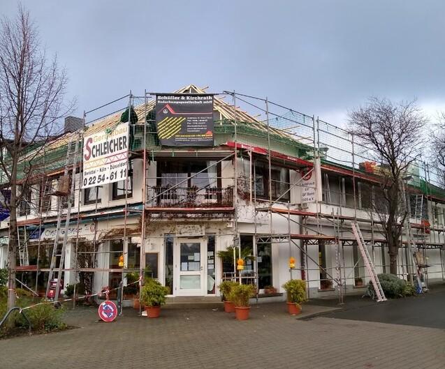 Geb&auml;ude mit Bauger&uuml;st und Werbebanner vor einer Fassadenrenovierung, umgeben von Pflanzen und B&auml;umen.